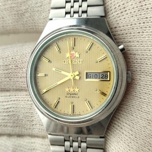 Op de afbeelding: Een zilverkleurig Orient horloge met een goudkleurige wijzerplaat en een roestvrijstalen band. De wijzerplaat toont de dag en datum. Het horloge wordt door een gehandschoende hand vastgehouden.