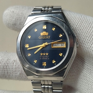 Op de afbeelding: Een zilverkleurig Orient horloge met een donkerblauwe wijzerplaat. Het horloge heeft goudkleurige wijzers en markeringen, een dag-datumweergave en een roestvrijstalen band. De wijzerplaat toont "Orient Crystal 21 Jewels".