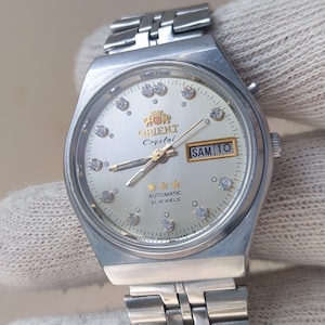 Puede incluir: Un reloj de pulsera automático Orient Crystal plateado con esfera clara. El reloj presenta una pantalla de fecha, marcadores de hora con forma de diamante y el texto "Orient Crystal Automatic 21 Jewels". El reloj es sujetado por una mano enguantada.