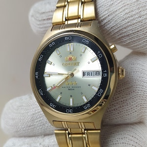 Op de afbeelding: Een goudkleurig Orient-polshorloge met een champagnekleurige wijzerplaat met goudkleurige uurmarkeringen en wijzers. Het horloge heeft een dag-datumvenster op de drie-uurpositie, een zwarte buitenring met minuutmarkeringen en een goudkleurige metalen schakelband.
