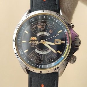 May include: An Orient automatic wristwatch with a black dial, gold-tone hands, and a date window. The watch features a multi-year calendar display, a silver-tone bezel with compass markings, and a black leather strap with orange perforated accents.
