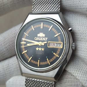 Puede incluir: Reloj Orient plateado con correa de malla. La esfera azul oscuro presenta agujas y marcadores dorados, con una visualización del día y la fecha. El reloj está sujeto por una mano enguantada. La esfera mide aproximadamente 3,5 cm de diámetro.
