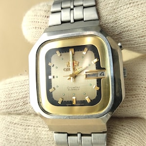 May include: An Orient Crystal watch with a square silver-tone stainless steel case and link bracelet. The dial features a gold-tone center with a black border, gold-tone hour markers, gold hands, and a day-date window at the three o'clock position.
