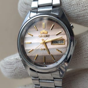 Op de afbeelding: Een zilverkleurig roestvrijstalen Orient-horloge met een metallic verloopwijzerplaat met horizontale strepen, goudkleurige uurmarkeringen en wijzers, en een dag-datumvenster op de drie-uurpositie met de tekst DON 25.