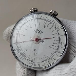 Pode incluir: Uma régua de cálculo circular vintage com mostrador branco, marcações pretas e vermelhas e uma moldura de metal prateado. A régua de cálculo tem dois botões na parte superior e a palavra "MATKA" está impressa.