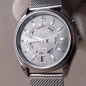 Può includere: Un orologio da polso argentato con un cinturino a maglia. Il quadrante dell'orologio è argentato con segni e lancette nere. Il quadrante include il marchio "ORIENT" e le parole "AUTOMATIC", "CRYSTAL" e "WATER RESIST 50m". La data è visualizzata in basso.