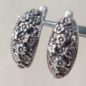 May include: A pair of silver-toned oval earrings. Each earring is densely covered with small, detailed floral designs. The earrings are held by a silver metal tool against a neutral background.