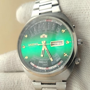 May include: A stainless steel Orient wristwatch featuring a vibrant green gradient dial with a perpetual calendar display. The watch includes gold-tone hour markers, luminous hands, a day-date window at the three o'clock position, and a metal link band.