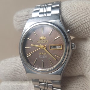 May include: An Orient Crystal watch with a metallic brown dial, gold-tone hands, and hour markers. The watch features a stainless steel case and link bracelet, with a day-date window at the three o'clock position displaying the text SAM 19.
