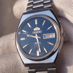 May include: Silver-toned Orient AAA Crystal 21 Jewels wristwatch with a dark blue face. The watch features gold-colored hands and markers, a day-date display, and a stainless steel band. The watch is displayed against a white background.