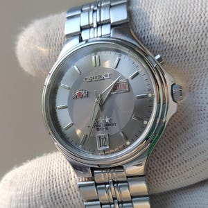 以下が含まれることがあります： メタリックグレーの文字盤を備えたシルバーカラーのオリエント製腕時計。3時の位置に曜日と日付の表示窓があり、シルバーのインデックスとステンレススチール製のリンクブレスレットが特徴です。文字盤にはオリエントのロゴとテキストが記されています。
