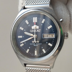 May include: A silver-toned Orient watch with a dark blue face and a mesh band. The watch face displays the day and date, along with the Orient logo and the words "WATER RESIST 50m". The watch is held by a gloved hand.