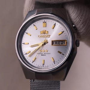 Puede incluir: Reloj Orient plateado con esfera plateada clara, manecillas y marcadores dorados. El reloj muestra el día y la fecha, con la fecha que indica "FRI 21". El reloj tiene una correa de malla y el texto "WATER RESIST 50m".