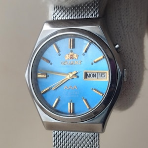 May include: A silver-tone wristwatch with a bright blue dial, gold-colored hour markers, and gold hands. The watch face features the Orient logo, the text AAA, and a day-date window displaying MON 15. It has a stainless steel mesh strap.