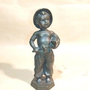 Puede incluir: Estatua de color bronce de un niño pequeño con sombrero y herramientas. El niño está representado con pantalones holgados y se encuentra sobre una base hexagonal. La estatua tiene un aspecto envejecido, lo que sugiere que es una pieza antigua o vintage.