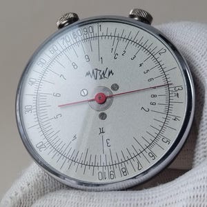 Pode incluir: Uma régua de cálculo circular vintage com uma caixa de metal prateado e um mostrador branco. O mostrador apresenta marcações pretas, números e um ponteiro vermelho. Dois pequenos botões são visíveis na parte superior. A palavra "MISKA" está impressa.
