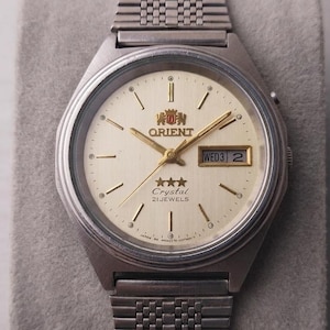 May include: A silver-toned Orient Crystal wristwatch with a cream-colored face. The watch features gold-colored hands and markers, a day-date display, and a stainless steel band. The watch face displays the Orient logo and the text "Crystal 21 Jewels."