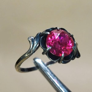 May include: A ring with a dark metal band and a single, round, bright pink gemstone.