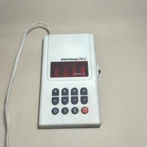 Puede incluir: Una calculadora electrónica blanca con una pantalla digital roja y un teclado con botones etiquetados '+', '-', 'O', 'C' y 'n'. La calculadora tiene el texto 'ЭЛЕКТРОННАЯ PB 0:' en la parte delantera.