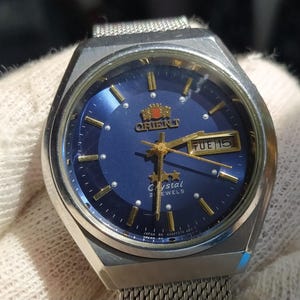 Op de afbeelding: Een zilverkleurig Orient horloge met een diepblauwe ronde wijzerplaat, goudkleurige uurmarkeringen en gouden wijzers. De wijzerplaat toont het Orient-logo, drie sterren, het woord Crystal en een dag-datumvenster met TUE 15. Mesh-horlogeband.