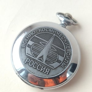 Op de afbeelding: Een zilverkleurig zakhorloge met een gedetailleerde wijzerplaat. De wijzerplaat heeft een cirkelvormig embleem met Cyrillische tekst en een centraal pijldesign. De tekst "РОССИЯ" is zichtbaar. Het horloge heeft een opwindstang.