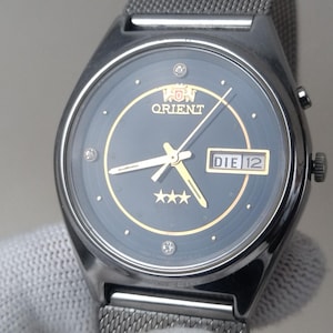 May include: A dark-faced Orient watch with a silver mesh band. The watch face has gold accents, including the Orient logo, hands, and three stars. The date display reads "DIE 12".