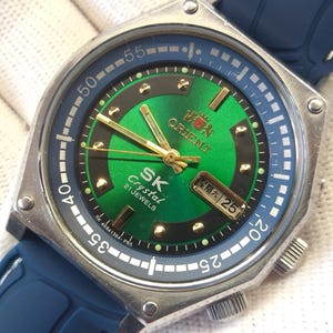 Op de afbeelding: Zilverkleurig Orient SK Crystal 21 Jewels horloge met groene wijzerplaat en gouden wijzers. Het horloge heeft een blauwe lunette en een blauwe rubberen band. De datum wordt weergegeven als zaterdag 25.