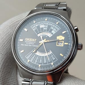 Peut inclure: Montre-bracelet automatique Orient argentée avec un cadran gris foncé. La montre dispose d'un calendrier pluriannuel, d'affichages du jour et de la date, et d'aiguilles et d'index dorés. Le bracelet est également argenté.