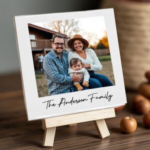 May include: A white framed photograph of a family of three, displayed on a small wooden easel. The photo shows a man, a woman, and a baby. The text "The Anderson Family" is written in cursive at the bottom of the photo.