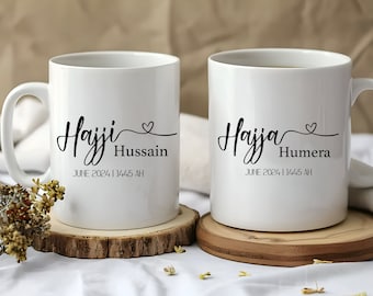 May include: Two white ceramic mugs with black text and a small heart design. The text on the mugs reads "Hajji Hussain June 2024 | 1445 AH" and "Hajja Humera June 2024 | 1445 AH".