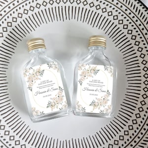 May include: Two clear glass bottles with gold lids and labels. The labels feature a floral design with white and gold accents. The text on the labels reads "A Gift of Zam Zam Water" and includes the names "Hussain & Sana" and the date "03.08.2025".