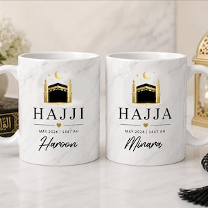 May include: Two white ceramic mugs with a marble effect. Each mug has a gold and black illustration of a building, the word "HAJJI" or "HAJJA", a name, and the date "MAY 2026 | 1447 AH".