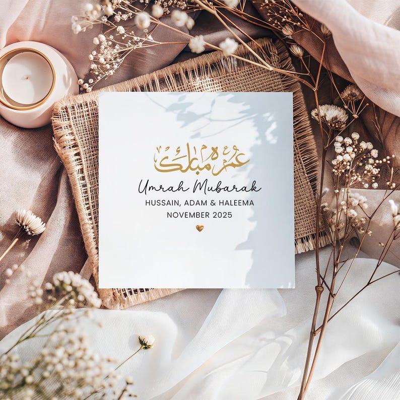 May include: A square white greeting card featuring gold Arabic calligraphy at the top, followed by the black text Umrah Mubarak, the names Hussain, Adam & Haleema, the date November 2025, and a small gold heart icon centered at the bottom.