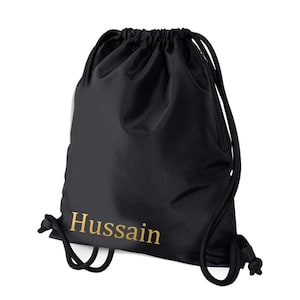 May include: Black drawstring bag featuring the name "Hussain" in gold. The bag has a drawstring closure and a zipped side pocket. The material is smooth and matte.