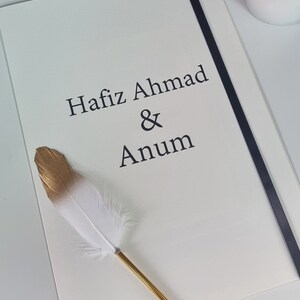 May include: A white card with black text that reads "Hafiz Ahmad & Anum". A gold and white feather pen is lying on the card.