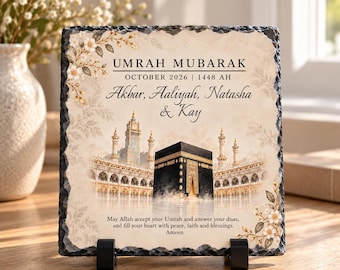 May include: A square slate plaque with rough edges featuring a printed illustration of the Kaaba in Mecca. The text reads UMRAH MUBARAK, OCTOBER 2026, 1448 AH, Akbar, Aaliyah, Natasha & Kay, and a prayer. It is supported by two black plastic stands.