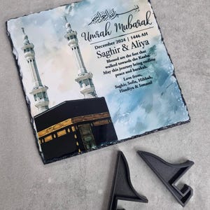 May include: A square slate plaque featuring a printed illustration of the Kaaba and two minarets against a blue watercolor background. The text reads Umrah Mubarak, December 2024, 1446 AH, Saghir & Aliya, and a personalized message. Two black plastic stands are included.