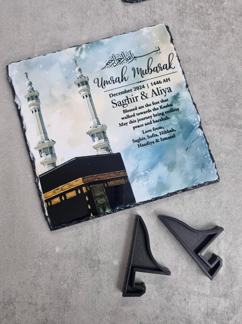 May include: A square slate plaque featuring a printed illustration of the Kaaba and two minarets against a blue watercolor background. The text reads Umrah Mubarak, December 2024, 1446 AH, Saghir & Aliya, and a personalized message. Two black plastic stands are included.