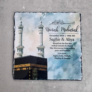 May include: A square slate plaque featuring a printed illustration of the Kaaba and two minarets. The text reads Umrah Mubarak, December 2024, 1446 AH, Saghir & Aliya, and includes a blessing message followed by the names Saghir, Sofia, Hibbah, Haadiya & Ismaeel.