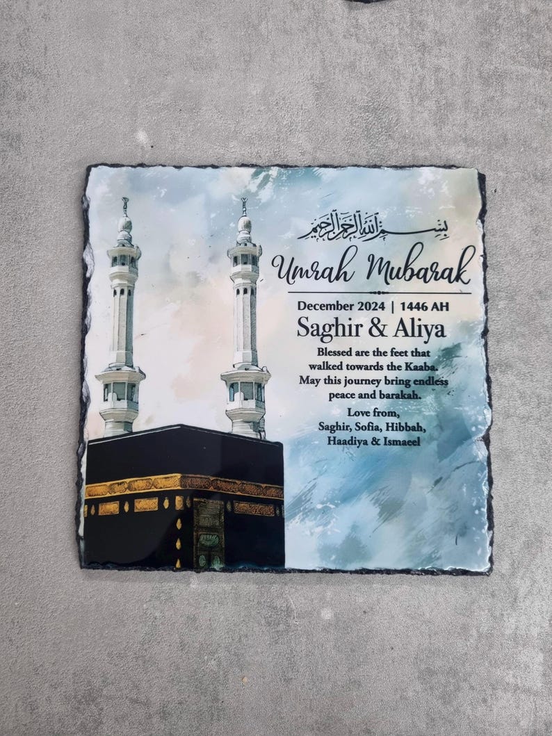 May include: A square slate plaque featuring a printed illustration of the Kaaba and two minarets. The text reads Umrah Mubarak, December 2024, 1446 AH, Saghir & Aliya, and includes a blessing message followed by the names Saghir, Sofia, Hibbah, Haadiya & Ismaeel.
