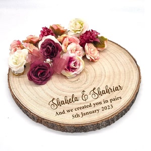 May include: A circular wooden slice adorned with an assortment of artificial flowers in pink, burgundy, and cream hues. The wood features engraved text: "Shahela & Shahriar" and "And we created you in pairs 5th January 2023."