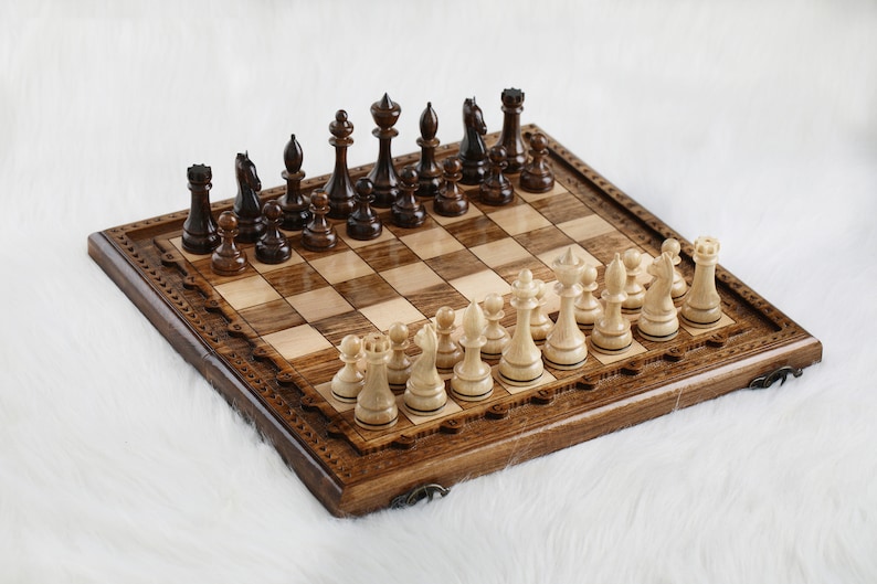 May include: A wooden chessboard with a carved border and a set of black and white chess pieces. The board is made of light and dark wood and has a traditional design.
