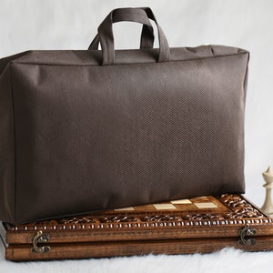 May include: A brown canvas carrying case with a zipper closure and a handle. The case is sitting on top of a wooden chessboard with carved details. There are three chess pieces on the right side of the image.