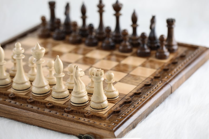May include: A wooden chessboard with a brown and beige checkered pattern. The board has a carved wooden frame with a decorative design. White chess pieces are arranged on the board, with black pieces in the background.