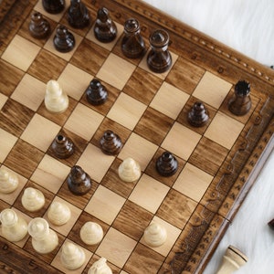 May include: A wooden chessboard with brown and light wood squares. The board is set up with black and white chess pieces. The pieces are arranged in a mid-game position. The board is on a white surface.