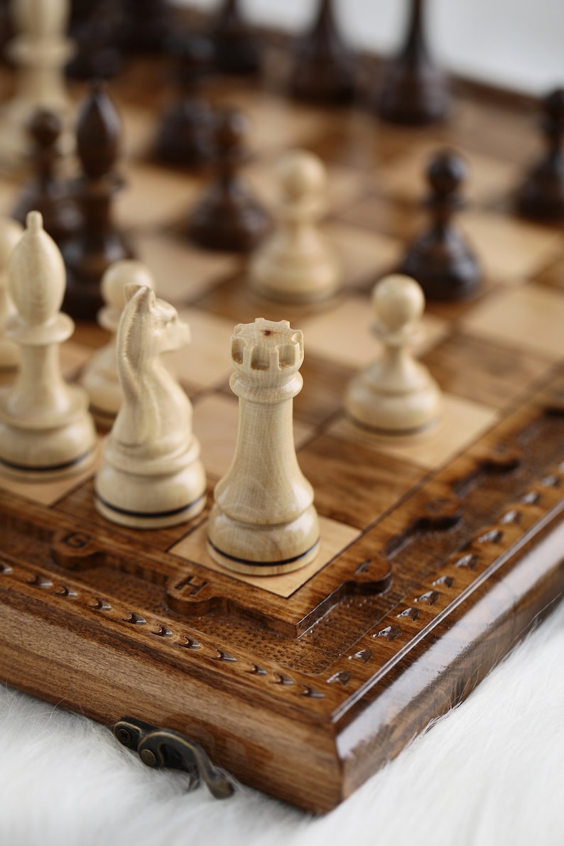 May include: A wooden chessboard with a carved design and a set of white chess pieces, including a rook, a knight, and a pawn. The chessboard is closed and has a brass latch.