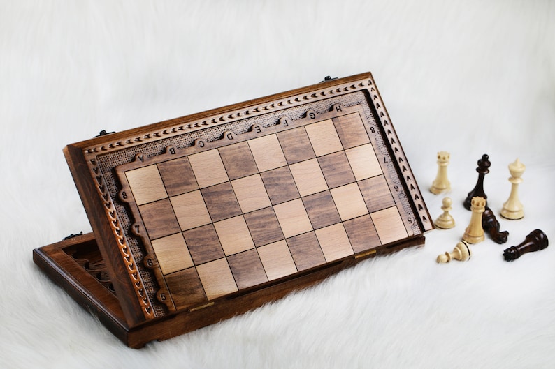 May include: A wooden chessboard with a carved design on the side. The board is open and the pieces are scattered around it. The pieces are made of wood and are painted in black and white.
