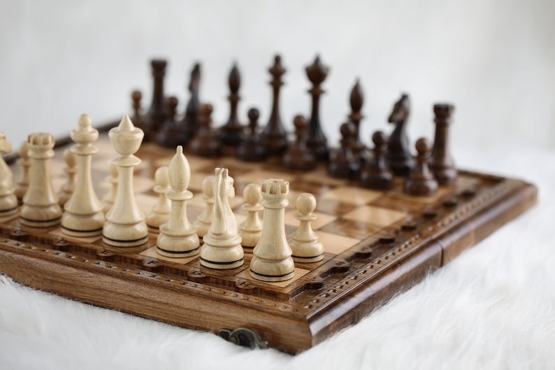 May include: A wooden chessboard with a carved design around the edge. The board is open and shows a game in progress with white pieces on the left and black pieces on the right.