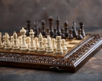 Könnte beinhalten: Ein Schachbrett aus Holz mit weißen und schwarzen Figuren, die für ein Spiel aufgestellt sind. Das Brett hat einen geschnitzten Holzrahmen mit aufwendigen Details.