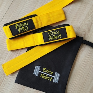 May include: Two yellow lifting straps with black stitching and a black drawstring bag. The straps have black patches with white text that reads "Bikini PRO" and "Erica Albert". The bag has a white barbell graphic and the text "Erica Albert" in yellow.